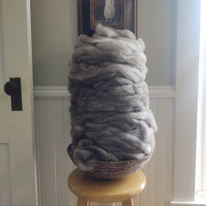 May include: A large amount of gray wool fiber is piled in a woven basket. The basket is sitting on a wooden stool. A framed picture of a white sheep is hanging on the wall behind the wool.
