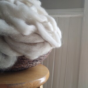 May include: A large amount of white wool fiber is piled in a brown wicker basket. The basket is sitting on a wooden stool.
