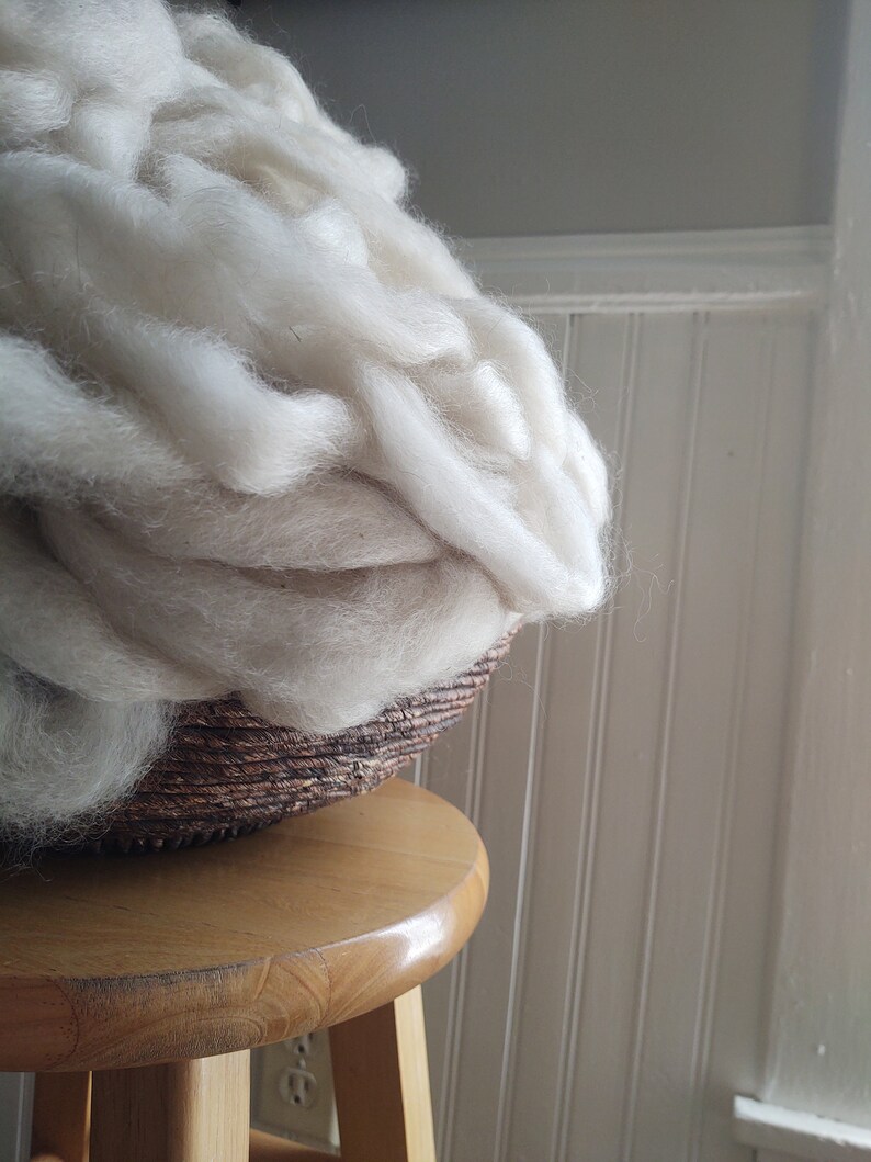 May include: A large amount of white wool fiber is piled in a brown wicker basket. The basket is sitting on a wooden stool.