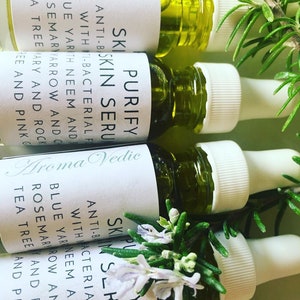 May include: Four small glass bottles with dropper tops filled with a green liquid. The bottles have white labels with the text "Purify Skin Serum" and "Aroma Vedic". The bottles are surrounded by sprigs of rosemary.