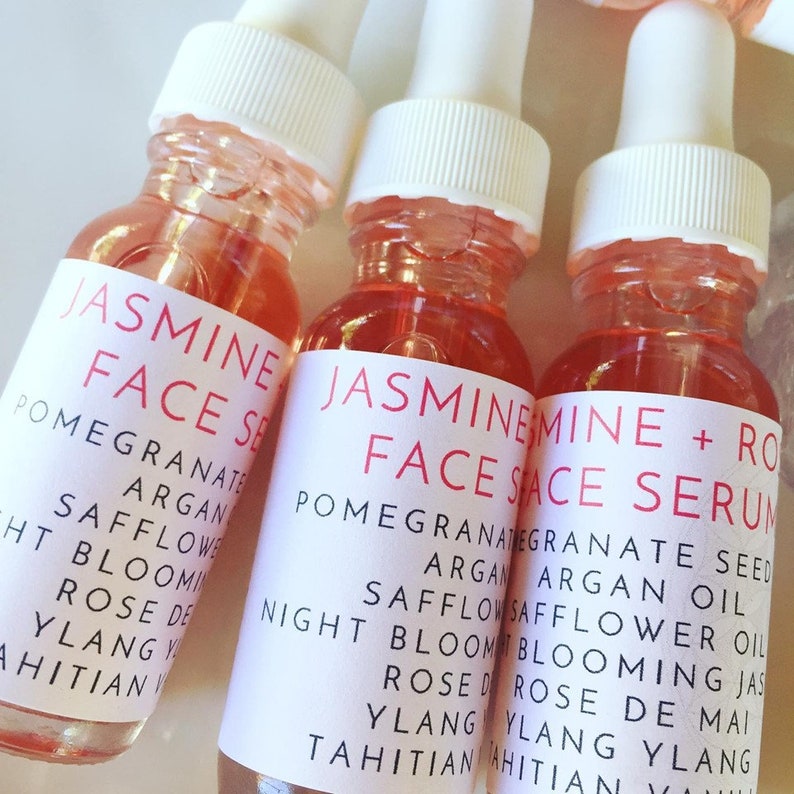 Op de afbeelding: Drie glazen flesjes met druppelaars gevuld met een roze vloeistof. De etiketten zijn wit met zwarte tekst die "Jasmijn Gezichts Serum", "Granaatappel Argan Saffloerolie", "Nachtblommende Jasmijn Roos de Mai Ylang Ylang Tahiti" luidt.