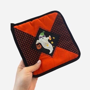 May include: A square quilted potholder featuring a central black diamond patch with an embroidered white ghost wearing a green hat and purple sunglasses, holding a candy corn and a jack-o'-lantern bucket, surrounded by orange and black polka-dot fabric.