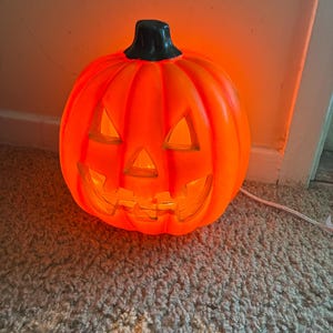 May include: A bright orange plastic jack-o'-lantern lamp with a black stem. The pumpkin features triangular eyes, a triangular nose, and a jagged mouth cutout, all illuminated from within by an internal light source, connected by a white cord.
