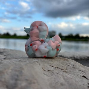 May include: A pink and blue rubber duck with a design of many smaller ducks inside. The duck is sitting on a gray rock.