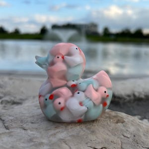 May include: A clear plastic duck figurine filled with smaller duck figurines in pink, white, and blue.
