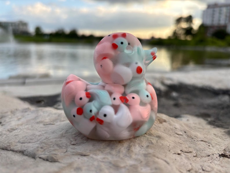 May include: A clear plastic duck figurine filled with smaller duck figurines in pink, white, and blue colors. The duck figurines have red beaks and eyes.