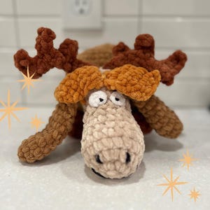 May include: A crocheted plush toy moose with brown and beige fur. The moose has large, round eyes and a long snout. The toy is lying on a white surface.