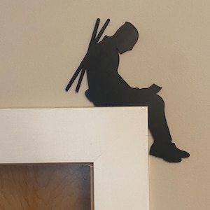 May include: A black metal silhouette of a person sitting and reading, with what appears to be two long objects on their back. The figure is perched on the corner of a white door frame, against a beige wall.