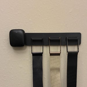 May include: A black metal belt rack with three belts hanging from it. The belts are black and beige. The rack has a square, black object on the left side.