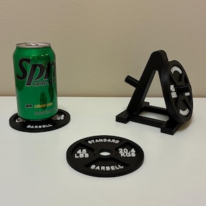May include: Black barbell weight plate coaster with the text "STANDARD", "45 LBS", and "BARBELL". A green can of soda sits on a matching coaster. A black weight rack and a weight plate are also present.