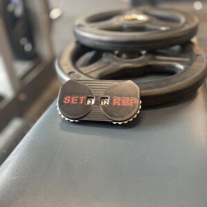 May include: A black and red workout counter with the words "SET" and "REP" on either side of a digital display showing "3" and "12".