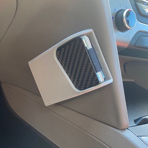 May include: A white 3D-printed car console organizer holding a carbon fiber wallet. The organizer is designed to fit in the car's interior, providing a convenient storage solution for small items. The wallet has a black and gray pattern.
