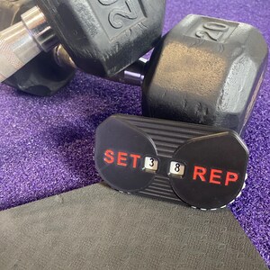 May include: Two black 20 pound dumbbells with a black and red counter that reads "SET 3 8 REP" on a purple mat.