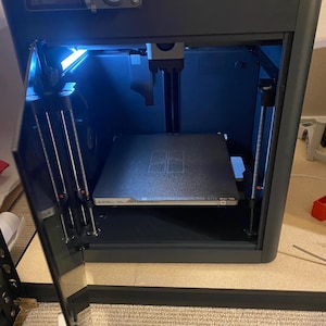 May include: A black 3D printer with a glass door open, revealing a blue light illuminating the print bed. The printer has a digital display and a control panel on the top. The printer is labeled "Bambu Lab P1S".