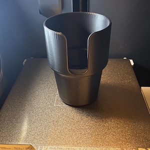 May include: A black 3D printed cup holder. The holder is designed to fit a standard-sized cup.