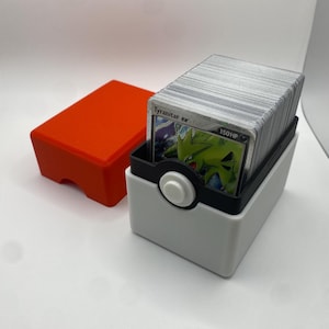 May include: A white and black Pokemon card deck box with a Tyranitar ex card visible. The box has a button on the front and a red deck box is in the background.