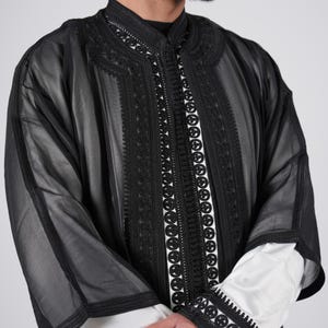Könnte beinhalten: Eine schwarze, durchsichtige Robe mit aufwendigen schwarzen Stickereien am Ausschnitt, an den Ärmeln und entlang der Vorderseite. Die Robe ist aus einem leichten Stoff gefertigt und hat eine lockere Passform.