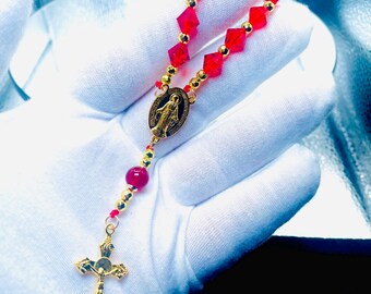 May include: A red beaded rosary with gold-coloured accents. The rosary features a gold-coloured crucifix and a gold-coloured oval pendant with a religious figure. The beads are faceted and diamond-shaped.