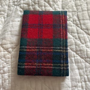 May include: A rectangular, folded wallet featuring a classic red and green plaid pattern. The design includes dark blue and yellow accent lines, creating a traditional tartan appearance. The wallet is made of a textured fabric.