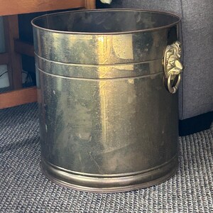 May include: A cylindrical brass bucket with a polished metallic finish. The container features a horizontal band detail around the upper section and a decorative lion head handle attachment on the side, which includes a circular ring pull.