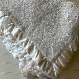 May include: A soft, light gray plush blanket featuring a thick, textured fleece surface on one side and a smooth, shiny white satin border that is gathered into ruffles along the edges, creating a layered and voluminous appearance throughout.