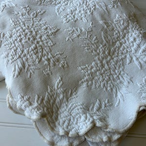 May include: A folded off-white bedspread featuring a raised, textured floral pattern across the surface. The fabric has a woven appearance with a scalloped edge finished with a light tan trim, creating a layered look with visible depth and detail.