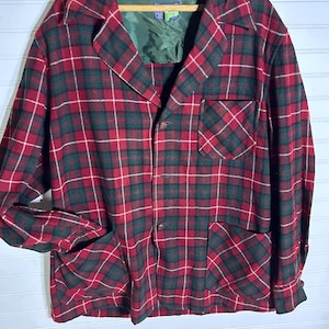 Op de afbeelding: Een rood, groen en wit geruit jack met een kraag, twee voorzakken en een borstzak. De jas heeft een knoopsluiting en hangt aan een houten hanger.