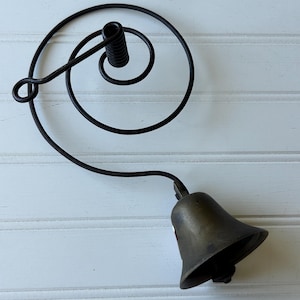 May include: A dark metal bell attached to a coiled wire spring mount. The bell has a tarnished brass finish with a flared opening. The mounting mechanism consists of a circular wire spiral that terminates in a small loop and a central coil.