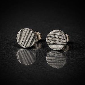 Può includere: Un paio di orecchini a lobo in argento. Ogni orecchino è un piccolo cerchio strutturato con una serie di linee parallele incise sulla superficie. Gli orecchini sono esposti su una superficie nera riflettente.
