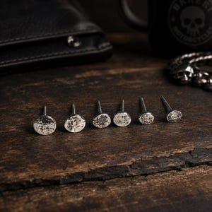 Könnte beinhalten: Sechs silberne Ohrstecker mit gehammerter Textur sind auf einer dunklen Holzoberfläche angeordnet. Die Ohrringe haben ein kreisförmiges Design. Eine schwarze Lederbrieftasche und eine schwarze Tasse mit Totenkopf-Design sind im Hintergrund.