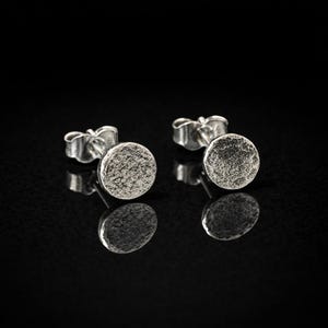 Può includere: Un paio di orecchini a lobo in argento con un design circolare strutturato. Gli orecchini sono esposti su una superficie nera riflettente, che ne evidenzia la forma e i dettagli. Hanno una chiusura a farfalla.