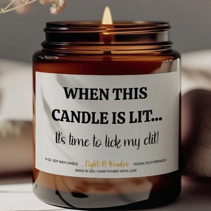 May include: A lit soy wax candle in a brown glass jar. The candle has a white label with the text "WHEN THIS CANDLE IS LIT... It's time to lick my clit!" The candle is made in the USA and is vegan and eco-friendly.