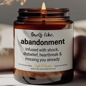 May include: A brown glass candle jar with a white label that reads "Smells like - abandonment" and "Infused with shock, disbelief, heartbreak & missing you already". The candle is lit and the label also includes the brand name "Light-It-Wonder", "9 oz Soy Wax Candle", "Vegan Eco-Friendly", and "Made in USA | Hand Poured with Love".