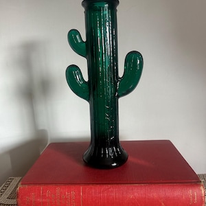 May include: A dark green glass vase shaped like a saguaro cactus with a cylindrical body, a flared base, and two upward-curving arms. The glass features vertical ribbed detailing along the stem and arms, creating a textured appearance.