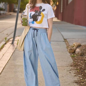 May include: A woman wearing a white crop top with a black cat and cactus graphic, paired with light blue wide-leg pants and white sneakers. She is holding a green drink in a glass with a straw and a beige tote bag.