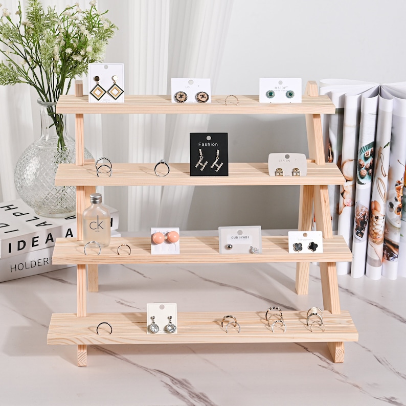 May include: A wooden tiered stand with four shelves for displaying jewelry. The stand is made of natural wood and has a rustic look. The stand is decorated with various earrings, rings, and necklaces.