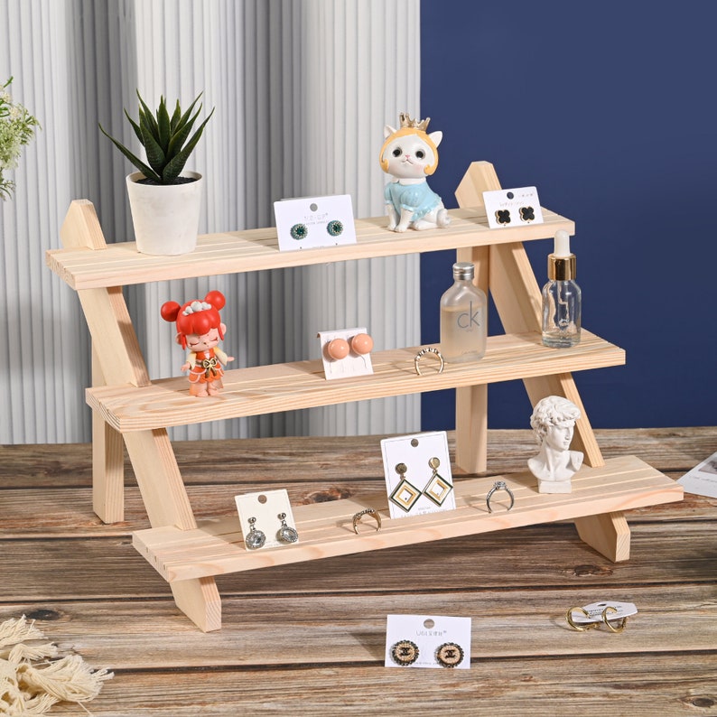May include: A three-tier wooden shelf display stand with a natural wood finish. The shelf is decorated with small figurines, jewelry, and other decorative items. The shelf is perfect for displaying small items in a retail setting or at home.