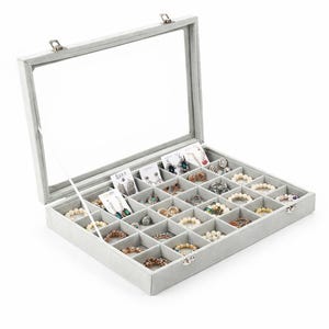 May include: A grey jewellery organiser with a clear glass lid, filled with various jewellery pieces. The organiser is divided into multiple compartments, showcasing bracelets, earrings, and rings. The interior is lined with a soft, grey fabric.