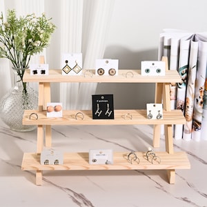 May include: A three-tier wooden stand with a natural finish, displaying various earrings and rings on small white cards. The stand is made of unfinished wood and has a rustic look. The stand is perfect for displaying jewelry or other small items.