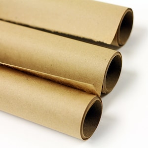 May include: Three rolls of kraft paper, each partially unrolled, revealing the interior. The paper is a warm brown color, with visible texture and slight variations in tone. The rolls are arranged diagonally against a white background.