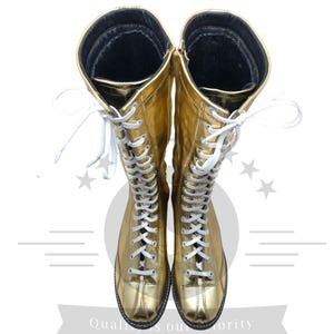 K&ouml;nnte beinhalten: Ein Paar goldfarbene, kniehohe Stiefel mit wei&szlig;en Schn&uuml;rsenkeln. Die Stiefel haben eine gl&auml;nzende, metallische Oberfl&auml;che und eine schwarze Sohle. Die Schn&uuml;rsenkel verlaufen &uuml;ber die Vorderseite der Stiefel, und die Oberseite der Stiefel hat ein schwarzes Innenfutter.