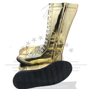 K&ouml;nnte beinhalten: Ein Paar goldfarbene, hoch geschnittene Stiefel mit wei&szlig;en Schn&uuml;rsenkeln und einem Rei&szlig;verschluss. Die Stiefel haben eine gl&auml;nzende, metallische Oberfl&auml;che und eine schwarze, strukturierte Sohle. Sportschuhe.