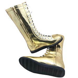 K&ouml;nnte beinhalten: Ein Paar goldfarbene, kniehohe Stiefel mit wei&szlig;en Schn&uuml;rsenkeln und schwarzen Sohlen. Die Stiefel haben einen Rei&szlig;verschluss an der Seite und bestehen aus einem gl&auml;nzenden, reflektierenden Material. Die Stiefel sind f&uuml;r Performance- oder Kost&uuml;mzwecke konzipiert.