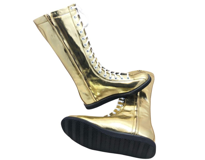 K&ouml;nnte beinhalten: Ein Paar goldfarbene, kniehohe Stiefel mit wei&szlig;en Schn&uuml;rsenkeln und schwarzen Sohlen. Die Stiefel haben einen Rei&szlig;verschluss an der Seite und bestehen aus einem gl&auml;nzenden, reflektierenden Material. Die Stiefel sind f&uuml;r Performance- oder Kost&uuml;mzwecke konzipiert.