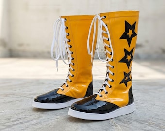 Puede incluir: Un par de botas altas con cordones que presentan una caña amarilla brillante con aplicaciones de estrellas negras y una puntera de charol negro. Las botas incluyen cordones blancos, una suela plana blanca y una silueta alta.