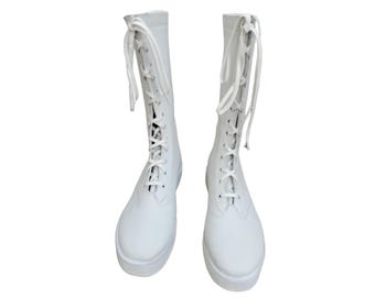 May include: A pair of white, knee-high boots with a lace-up design. The boots have a rounded toe and a thick sole. The laces are white and extend from the top of the boot to the toe. The boots are made of a smooth, solid white material.