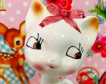 May include: White ceramic figurine of a cat with a pink bow, brown eyes, and a painted blush on its cheeks. The cat is sitting with its tail curled around its body.