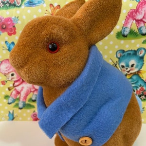 May include: A brown plush rabbit wearing a blue jacket with a button. The rabbit has red eyes and is sitting on a white surface.