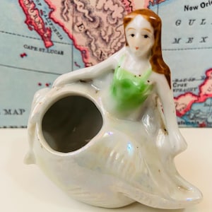 May include: A ceramic mermaid figurine in a seashell. The mermaid has brown hair and a green top. The seashell is white with iridescent highlights. The background includes a vintage map.