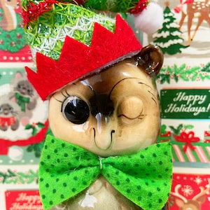May include: A ceramic owl figurine wearing a green and red elf hat with a white pom-pom and a green bow tie with white polka dots. The owl is winking and has large black eyes. The background is a Christmas-themed wrapping paper with a deer and the words "Happy Holidays!"